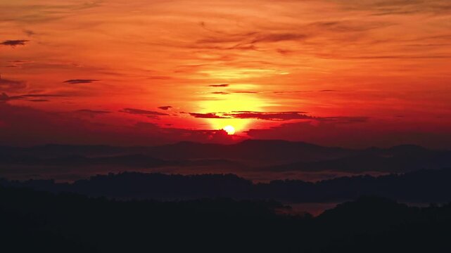 Cinematic timelapse of a fiery sunrise over mountain ridges. Vibrant orange and red hues fill the sky as the sun rises over a moving sea of clouds and silhouetted tropical peaks.