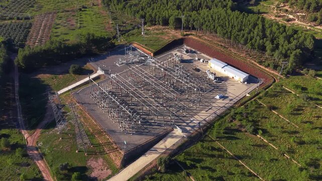 High voltage electrical substation surrounded by green pine forest hills, featuring transmission towers, power lines, transformers and switchgear viewed from above