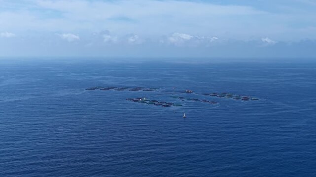Offshore fish farm with circular net cages floating on deep blue open ocean, scattered clouds on horizon, viewed from above showing aquaculture installation at sea