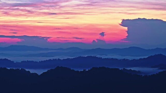 Cinematic dawn timelapse over mountain ridges. Vibrant pink and purple sky above a moving sea of clouds and silhouetted tropical peaks during the early morning transition.