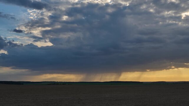 Timelapse of clouds moving over an agricultural field during sunset. A thunderstorm rain cloud with a visible rain band is floating across the sky. Heavy rain in the village in summer.