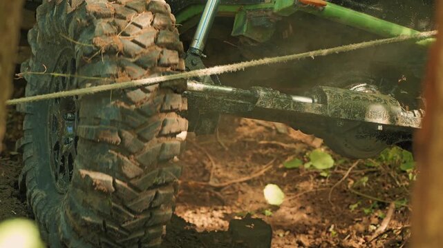 A close-up of a rugged SUV tire covered in mud. The vehicle's suspension and axle are visible during extreme off-road competition. The winch rope crosses the frame and catches on the tire