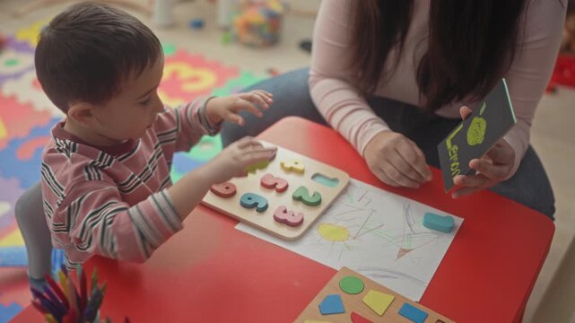 Boy reaches for wooden number puzzle on red table in studio while woman shows a flashcard and points; early learning concentration.