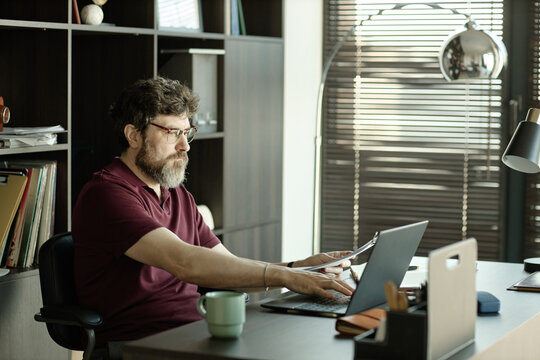 Middle aged Caucasian man working on laptop while reviewing documents at desk in home office, managing remote tasks and productivity
