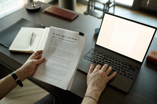 Close up of young adult man editing manuscript while comparing printed pages with laptop document at desk, illustrating content creation, publishing, copywriting marketing use
