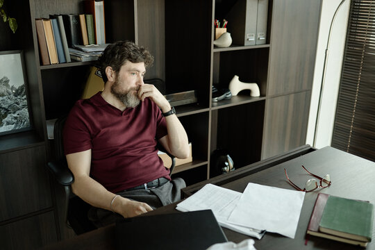 Middle aged Caucasian man sitting at office desk, thinking over pages of novel, suffering from creative block 
