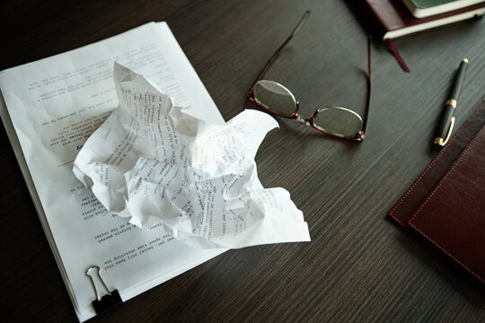 Close up of crumpled script pages resting on desk beside eyeglasses and pen, suggesting editing, rejection, or creative block. Useful for writing, publishing, productivity concepts
