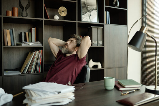 Middle aged Caucasian man leaning back at desk, stretching and thinking during paperwork review in home office, showing work fatigue and focus
