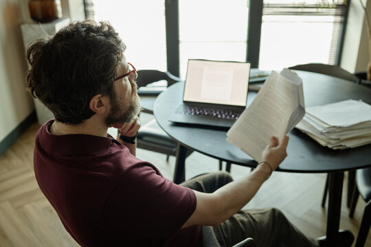 High angle view of Caucasian adult man reviewing printed novel beside laptop, working from home on paperwork
