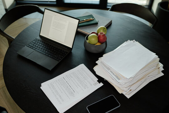 Wide angle shot of laptop displaying document beside paper stacks and smartphone on table
