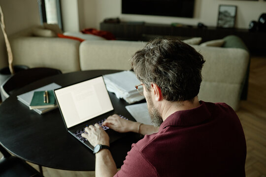 Middle aged Caucasian man working on laptop at home, managing documents and typing at table
