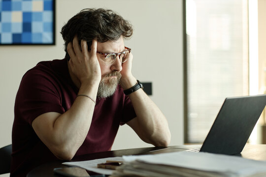 Middle aged Caucasian man holding head while staring at laptop, showing work stress and burnout during remote job as writer
