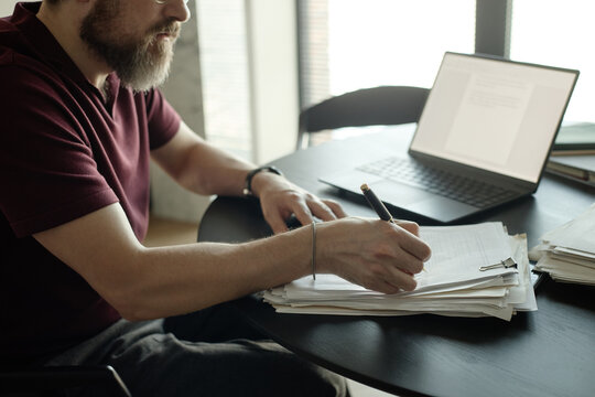Caucasian young adult man reviewing novel and writing notes beside laptop at desk,
