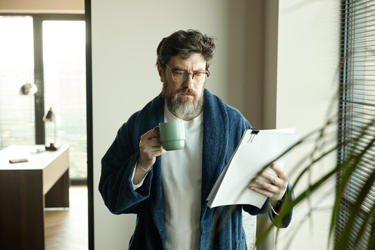 Middle aged Caucasian man reading pages of personal novel and drinking coffee at home, managing morning paperwork and routine
