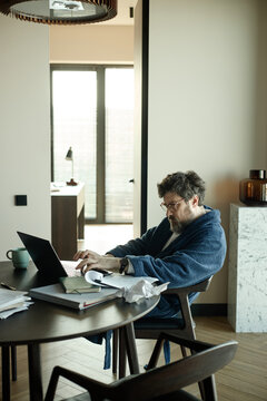 Vertical shot of middle aged Caucasian man working on laptop at home, typing and reviewing papers at table, representing remote work and productivity
