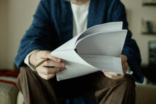 Unrecognizable man reviewing printed novel and holding pencil while sitting at home, suggesting decision making
