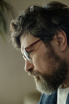 Vertical shot of middle aged Caucasian man wearing eyeglasses, looking downward and concentrating in profile, suggesting focused remote work or thoughtful decision making
