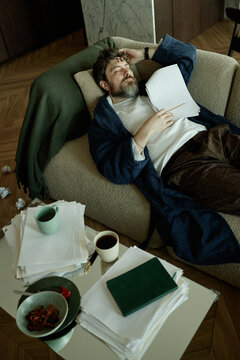 Tired author lying on comfortable sofa and sleeping, holding printed script 