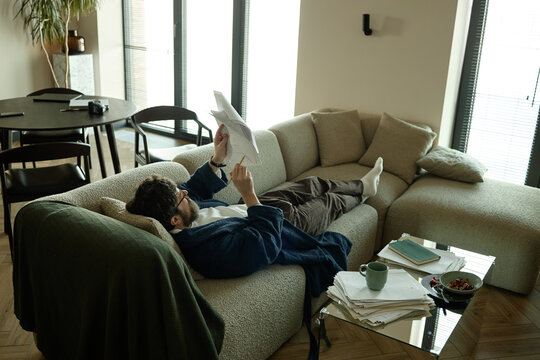 Wide shot of young adult Caucasian man reviewing paperwork while lying on sofa at home, reading and checking spelling
