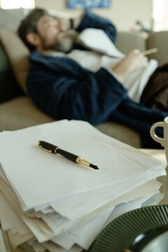 Vertical shot of middle aged Caucasian man resting on sofa while reviewing paperwork and holding pen, suggesting stress or fatigue
