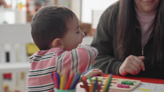 Boy reaches for wooden puzzle piece on table while adult guides hand in a building; concentration learning.