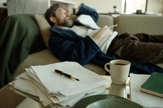 Middle aged Caucasian man reclining on sofa, reviewing paperwork and holding pen, suggesting stress or fatigue at home
