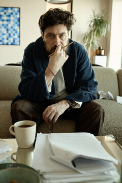 Portrait of Caucasian middle aged man sitting on sofa, thinking over novel, its chapters and holding pencil at home
