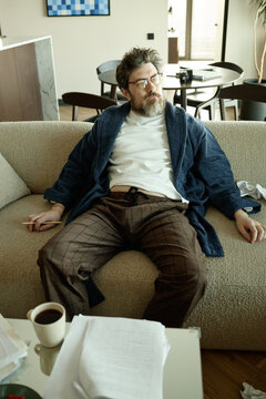 Middle aged Caucasian man sitting on sofa, holding pencil and looking away after paperwork, suggesting burnout or creative block
