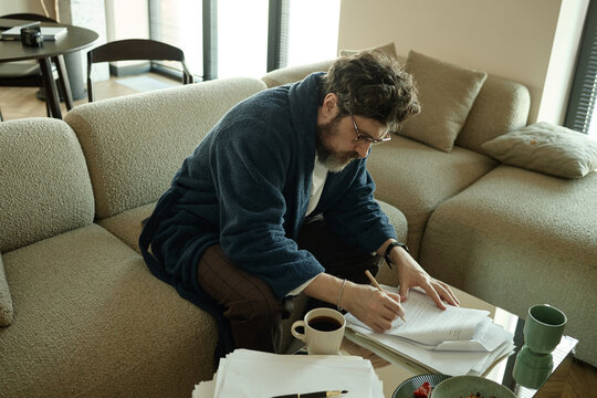 Caucasian middle aged man reviewing paperwork and writing notes on sofa at home while coming up with new plot twist

