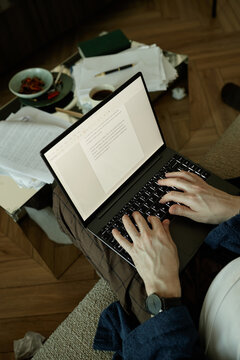 Vertical shot of unrecognizable author resting on sofa, holding laptop on laps while writing new chapter of novel 