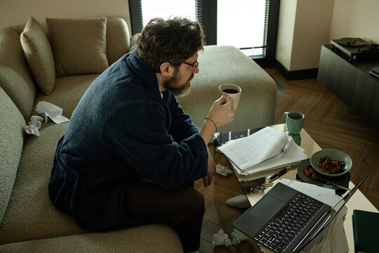 High angle view of middle aged Caucasian man drinking coffee while reviewing documents beside laptop at home, showing remote work stress

