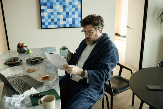 Middle aged Caucasian man wearing bathrobe reading new chapter of novel and drinking coffee beside laptop at home
