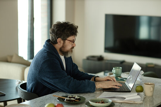 Caucasian man in bathrobe sitting at dining table and using laptop while working on new novel 