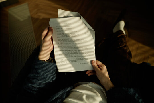 Overhead shot of young adult man holding printed manuscript and reviewing text at home, suggesting editing or script reading
