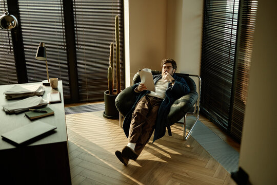 Wide angle view of middle aged Caucasian man reviewing novel and thinking in spacious work zone at home
