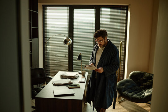 Wide shot of Caucasian adult man reviewing printed novel in home office dimly lit by sunrays
