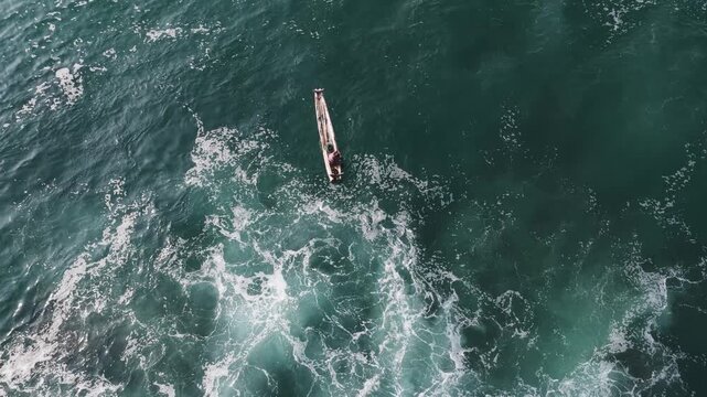 Aerial view of a lone fisherman in a small wooden boat navigating through turquoise ocean waves and white foam Kovalam, Kerala, India.
