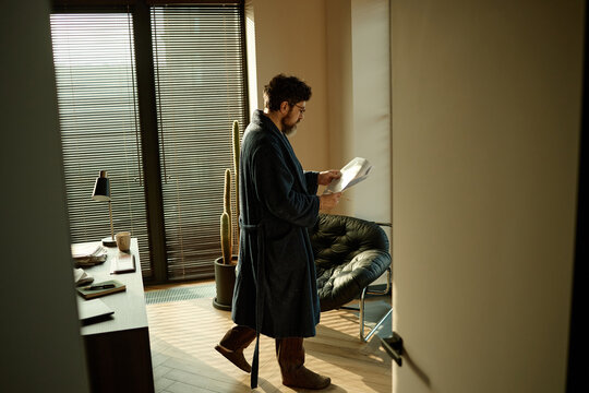 Middle aged author holding pages on novel while reading it, standing in center of home office 