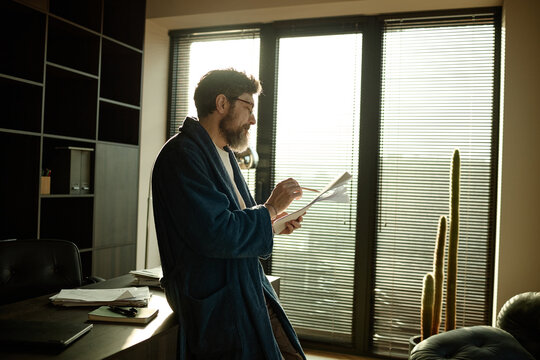 Middle aged author in deep blue bathrobe leaning on table and reading printed novel while working at home
