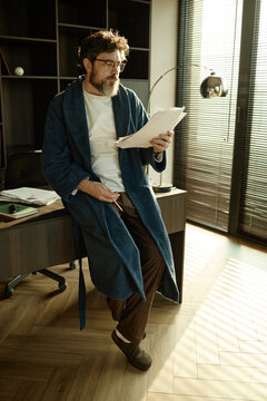 Vertical shot of middle aged Caucasian man in bathrobe reading script while leaning on desk in home office
