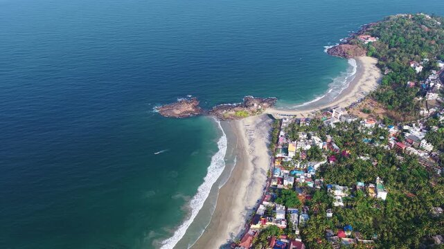 Aerial view of the sandy coastline and turquoise waters of Kovalam beach surrounded by lush palm trees and coastal buildings in Kovalam, Kerala, India.