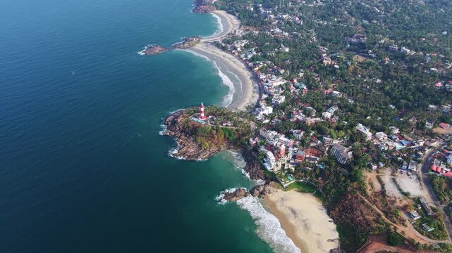 Aerial view of the red and white Vizhinjam Lighthouse on a rocky cliff overlooking the sandy beaches and turquoise waters of the Arabian Sea in Kovalam, Kerala, India.