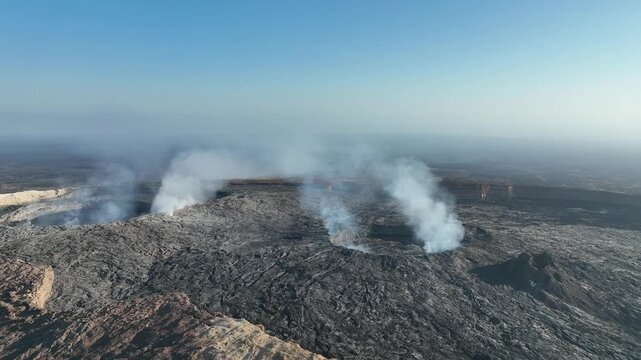Aerial view of smoke rising from volcanic craters in the Danakil Desert with vast rocky terrain under a clear sky Dalol, Afar, Ethiopia.