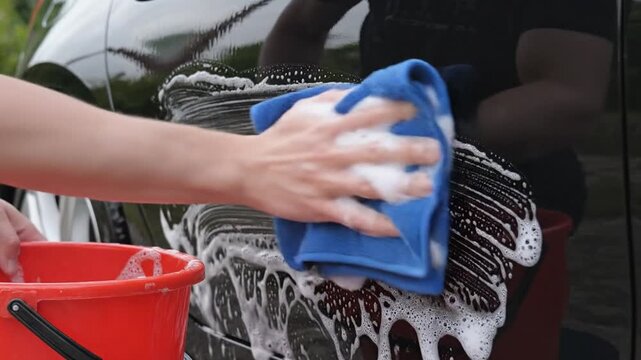 Hand washing black car door with blue cloth and orange bucket, foam and suds visible in outdoor setting