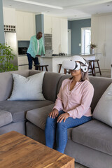 Obraz premium Couple relaxing at home, woman wearing pink blouse, VR headset on gray sofa, man using laptop