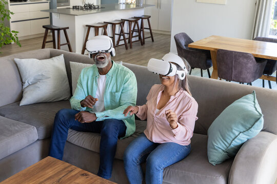 Couple laughing while wearing white VR headsets at home on gray sofa with coffee table
