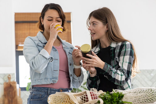 Diverse female friends unpacking groceries in kitchen, sniffing yellow jar and reading dark label