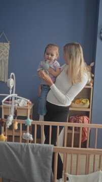 Woman holds toddler by a wooden crib, shows a plush toy and a pacifier while the child inspects it against a blue nursery wall; tenderness family bonding.