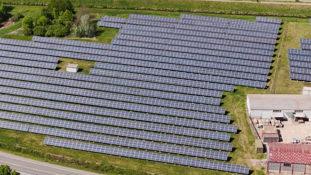 Aerial view of rows of photovoltaic solar panels installed on a green field near industrial buildings and lush trees in Slavkov u Brna, South Moravian Region, Czechia.