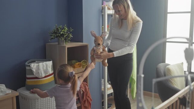 Woman handing a stuffed kangaroo to a little girl in a nursery inside a building, daughter reaches with outstretched hand; tenderness family bonding.